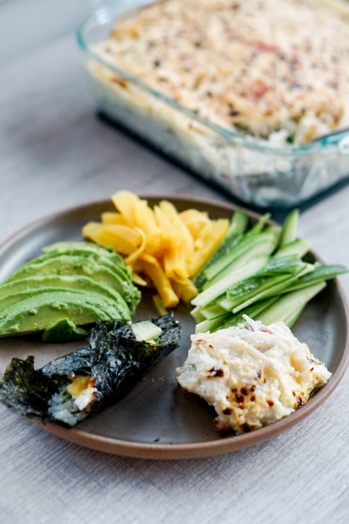 Plate of broiled sushi bake, cucumber, pickled daikon, avocado, and seaweed with casserole dish of sushi bake in the background.