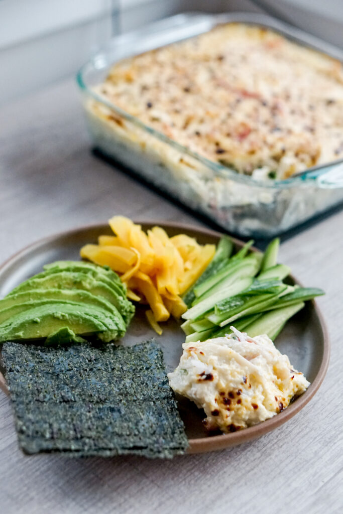 Plate of broiled sushi bake, cucumber, pickled daikon, avocado, and seaweed with casserole dish of sushi bake in the background.