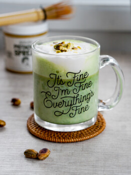 pistachio matcha latte front view in glass mug with matcha whisk and pistachio butter in background